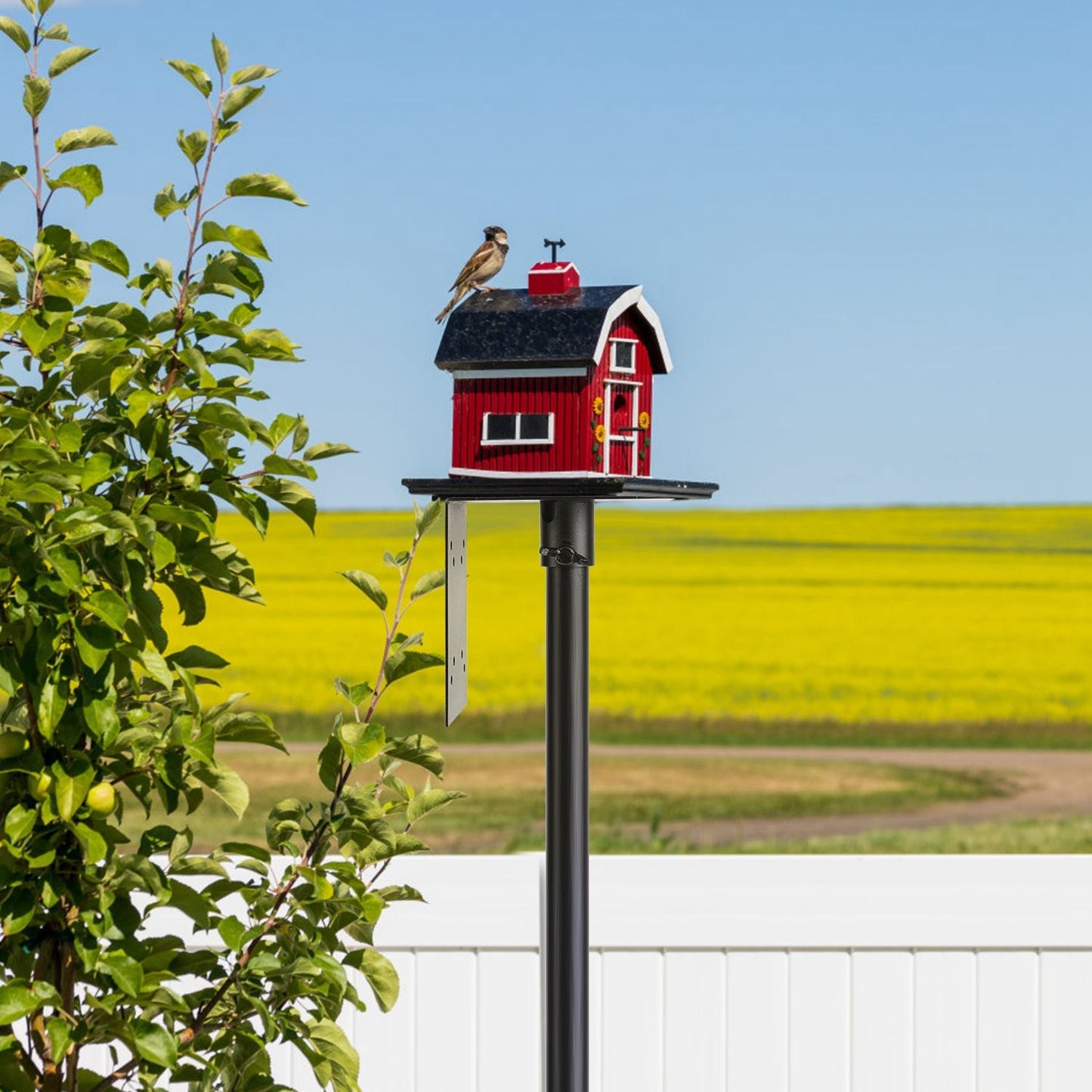 Short pole with red birdhouse on top panel standing in front of open field and blue sky