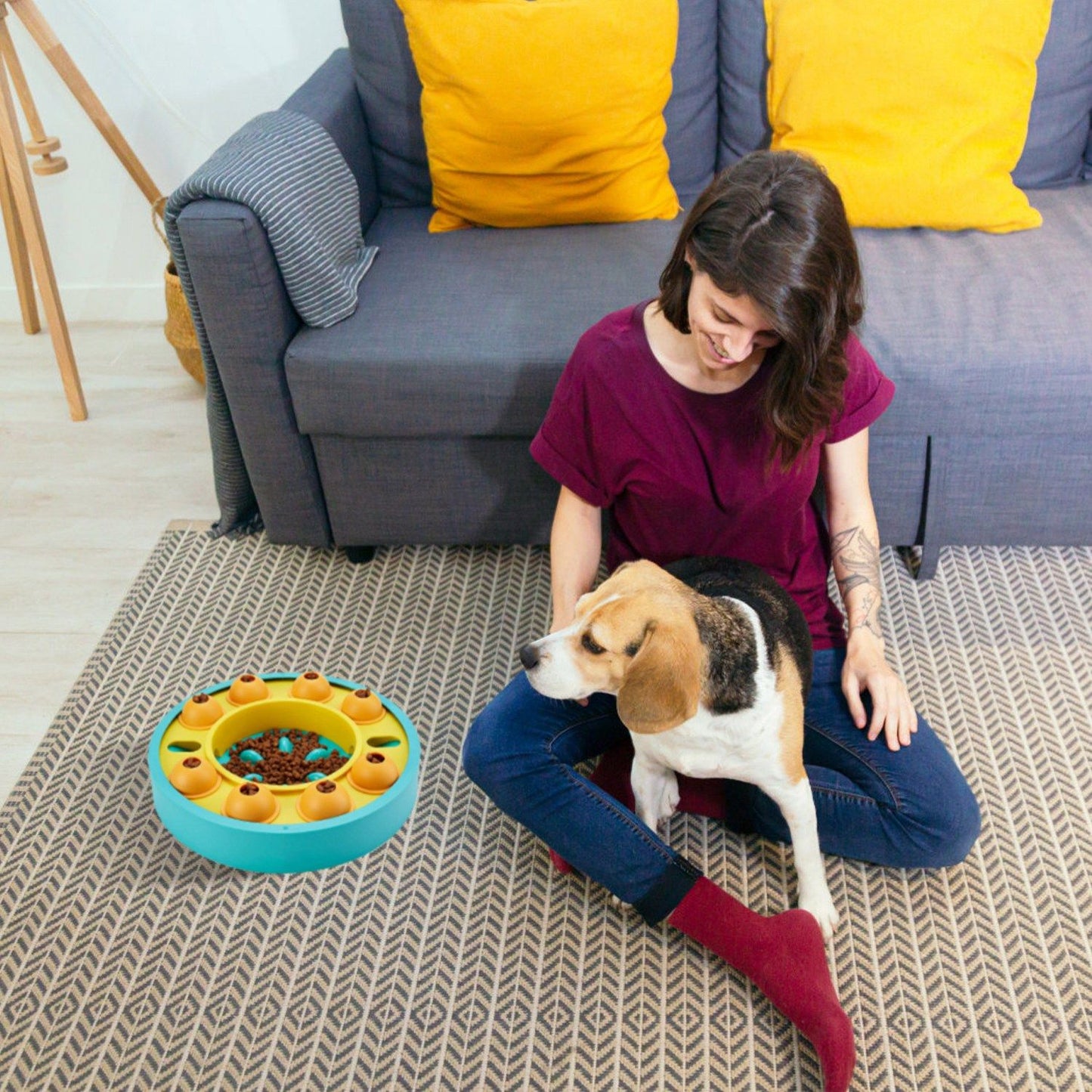 Person on floor with dog next to a blue puzzle feeder placed on carpet