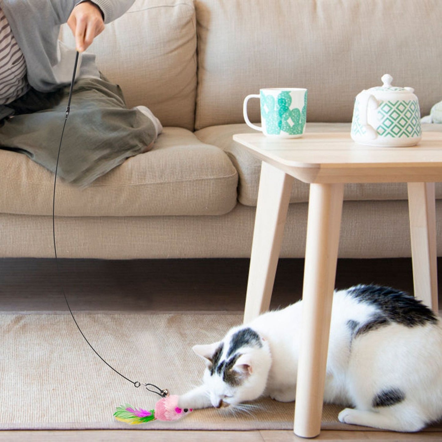 Person waving wand toy while cat bats a mouse attachment low near a coffee table