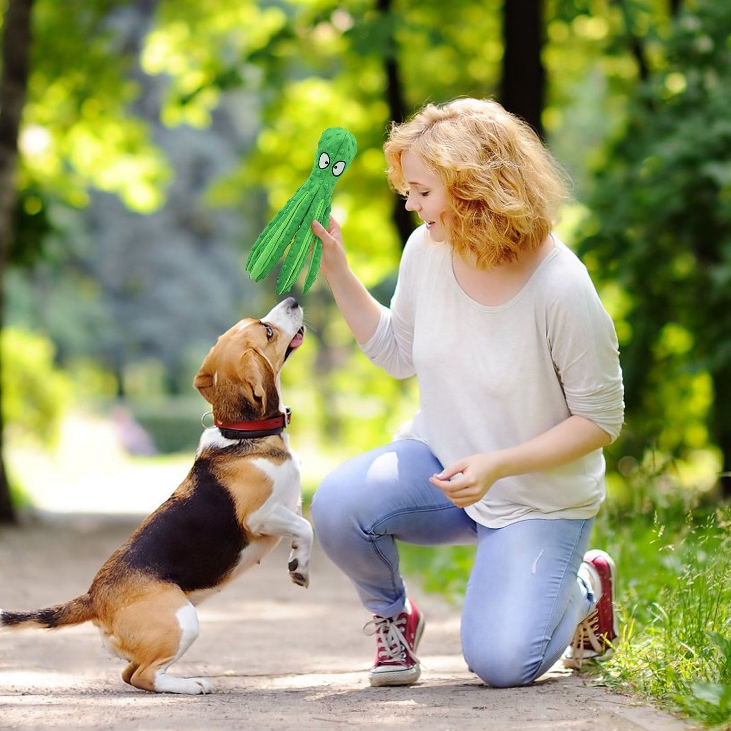 Person kneeling outdoors holding a green octopus toy as a beagle jumps to reach it