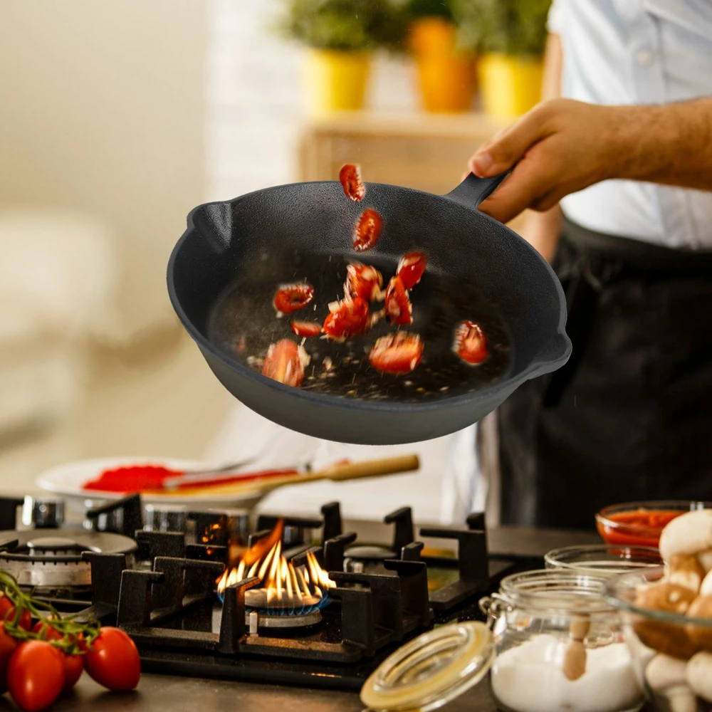 Person cooking in a modern kitchen, tossing fresh sliced tomatoes in a cast iron frying pan over a gas stove, surrounded by colorful ingredients and flames.
