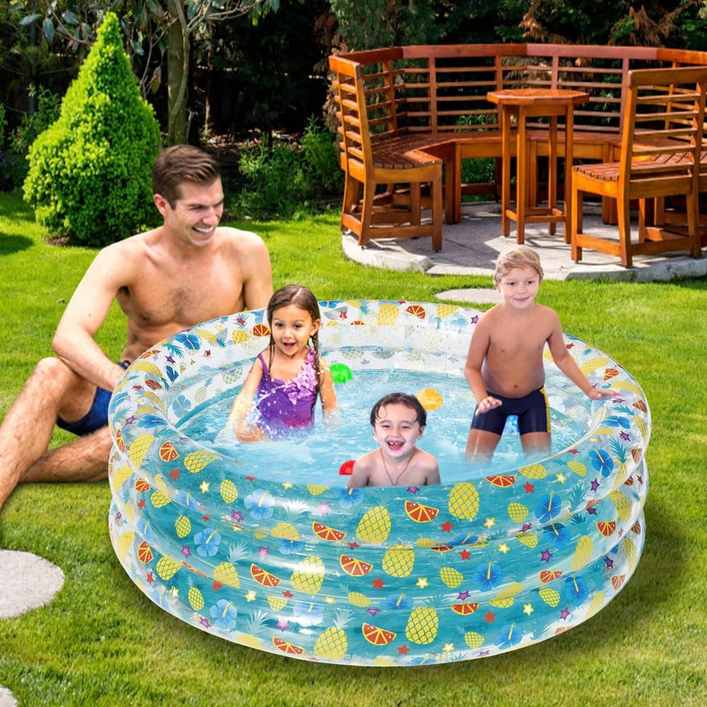 Inflatable kiddie pool with colorful fruit pattern, two children playing with toys, and an adult relaxing by the pool in a grassy backyard setting with wooden furniture in the background.