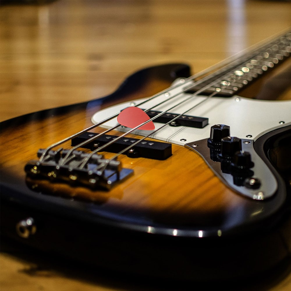 Close-up of a bass guitar with a red pick resting on the strings, showcasing its glossy finish and intricate controls against a warm wooden background.