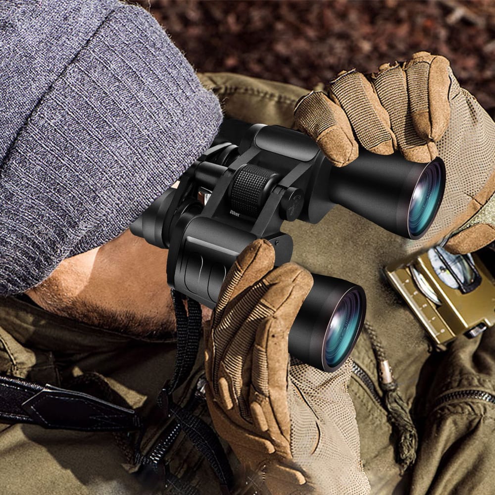 Man in tactical gloves using compact binoculars for outdoor observation, wearing a knit hat and camouflaged clothing.