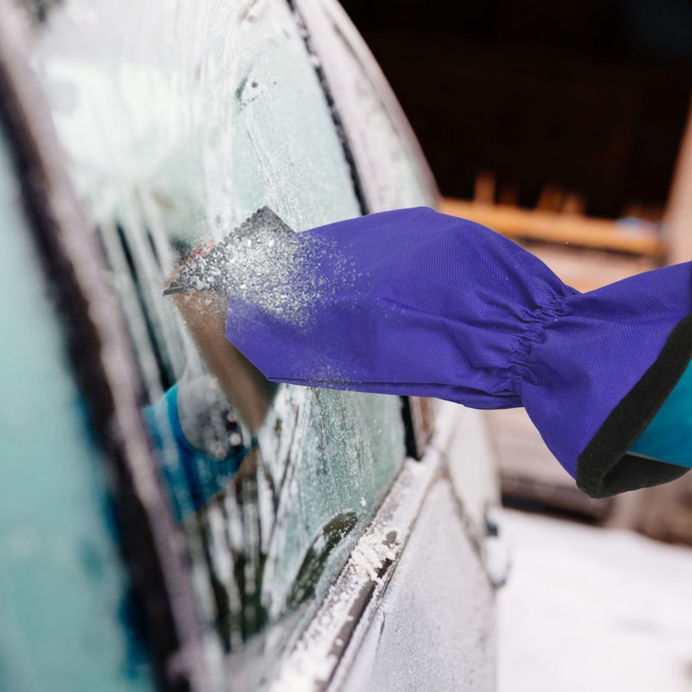 Person using a purple winter glove and ice scraper to clear snow and ice from a car window on a snowy day.