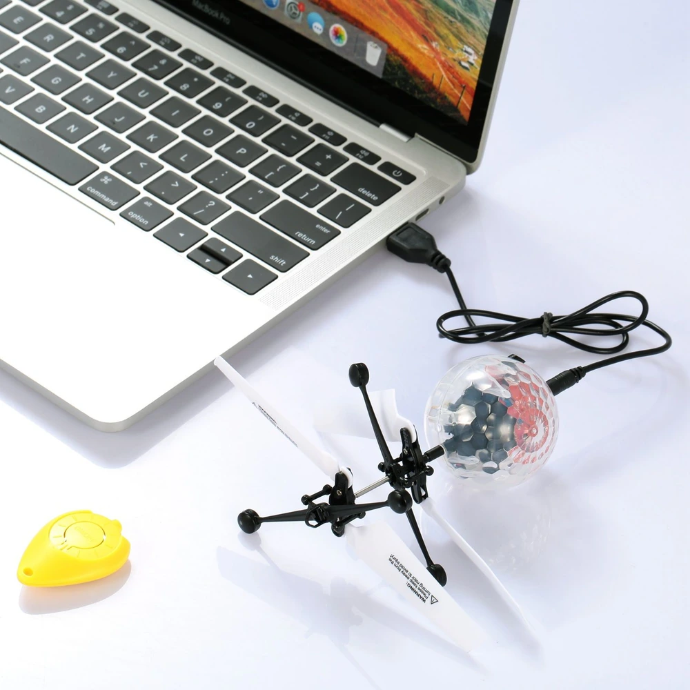 Mini remote-controlled helicopter charging via USB next to a laptop, featuring a yellow controller and a colorful LED light ball, perfect for indoor entertainment.