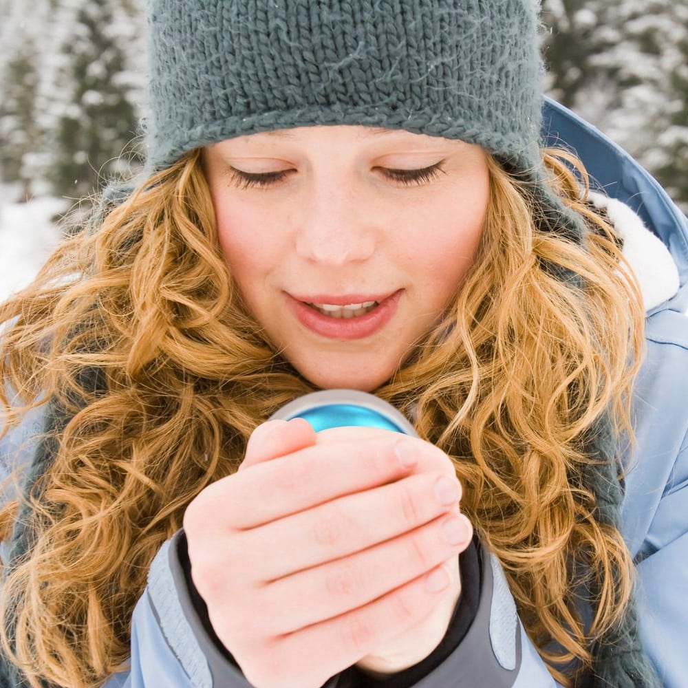 A woman with curly hair wearing a blue winter jacket and a knit hat holds a blue and white travel mug in a snowy outdoor setting, perfect for enjoying hot beverages in cold weather.