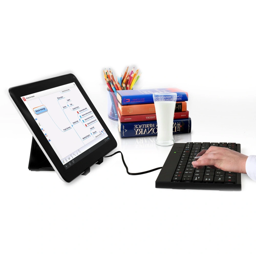 Person typing on a black keyboard connected to a tablet displaying a mind mapping application, with a stack of books and a glass of milk in the background.