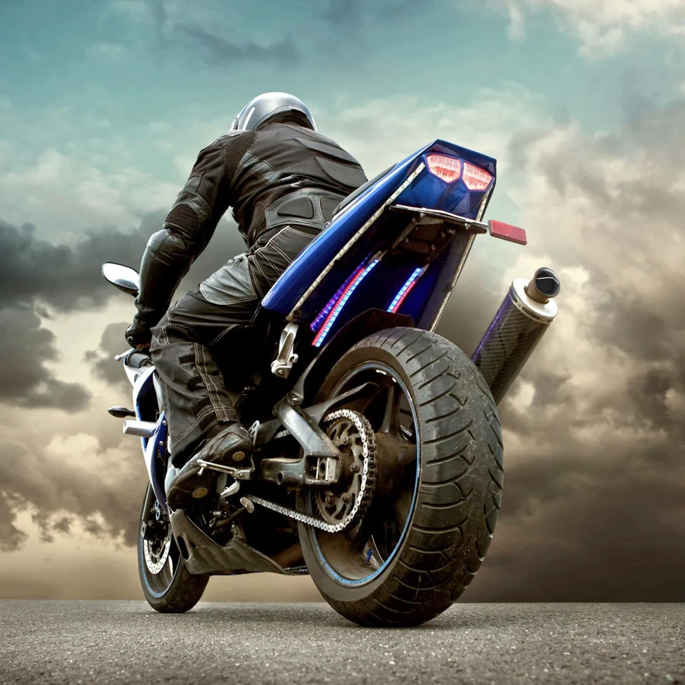 Sport motorcycle with a rider in black gear, showcasing sleek blue detailing and a powerful exhaust, set against a dramatic cloudy sky background.