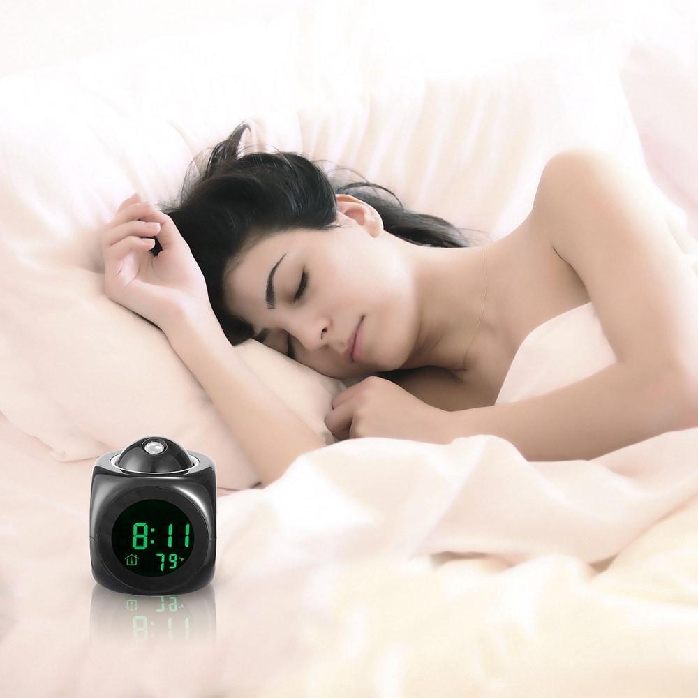 Digital alarm clock displaying time and temperature while a person rests peacefully in bed, surrounded by soft white bedding.