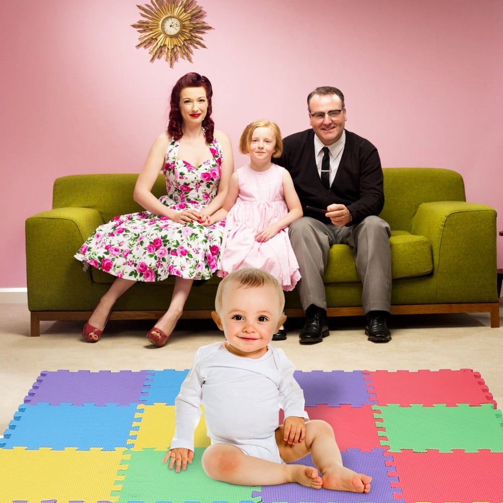 Family sitting together on a green couch in a brightly colored living room, with a baby in white clothing sitting on a multi-colored play mat in the foreground.