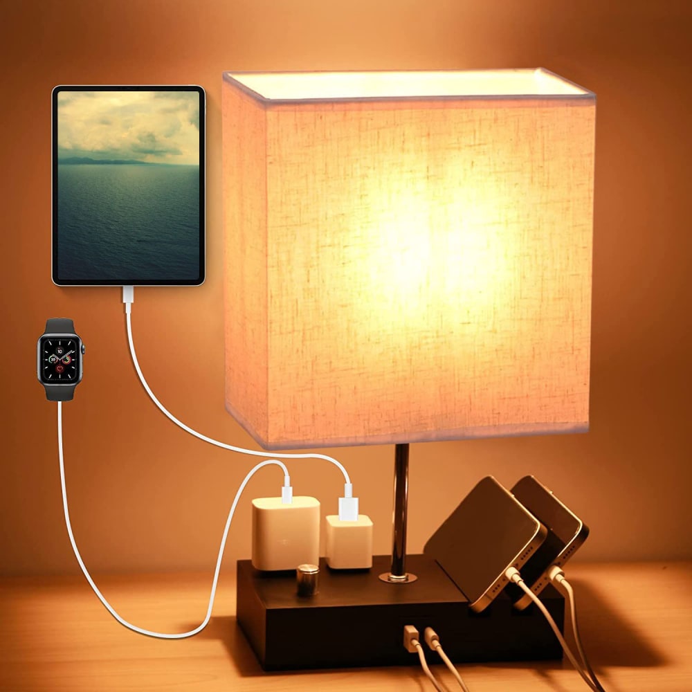 Stylish table lamp with warm light, charging a tablet and two smartphones, alongside a smartwatch, showcasing a modern, multi-device charging setup.