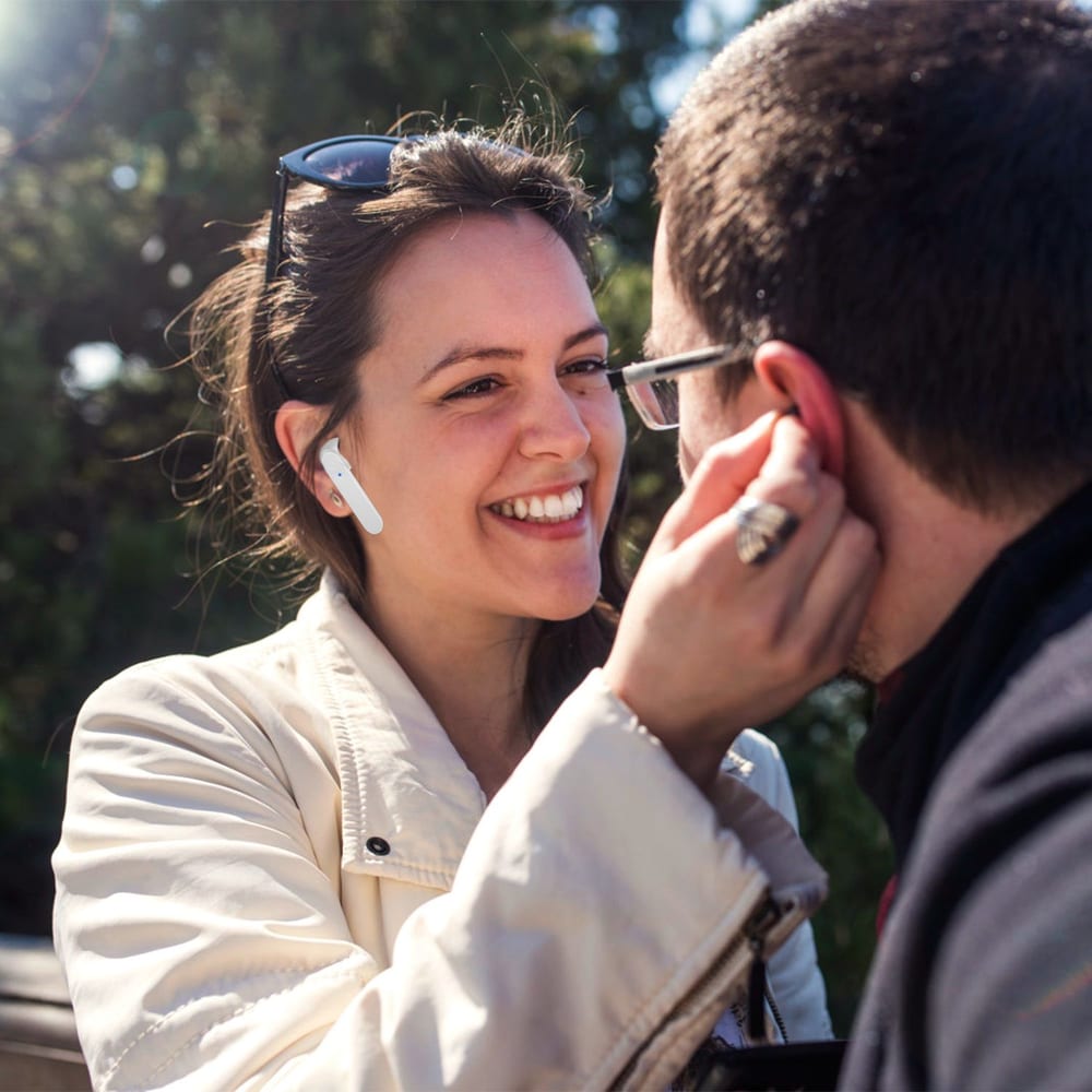 Two people interacting outdoors, a woman in a light jacket wearing wireless earbuds, and a man in glasses gently touching her ear, symbolizing a moment of connection or listening.