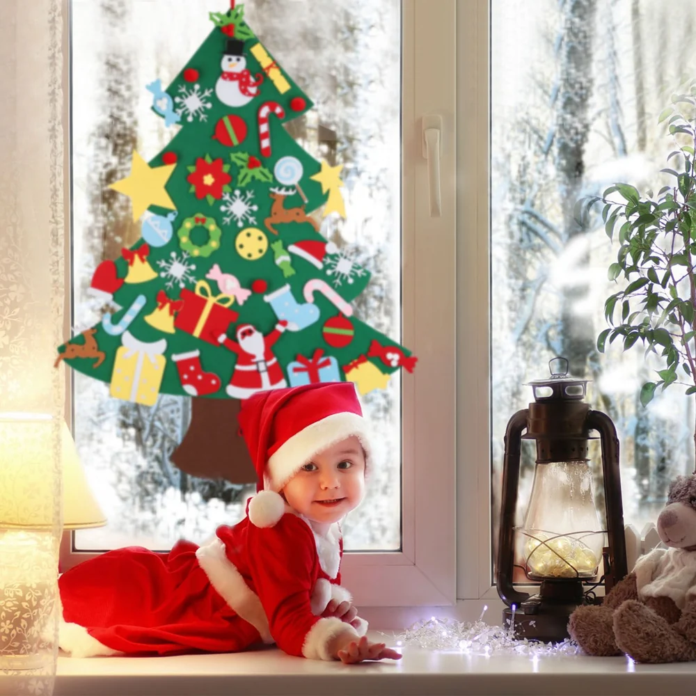 Cute baby in a Santa costume lying on a window sill next to a lantern and a teddy bear, with a festive Christmas tree decoration visible in the background.