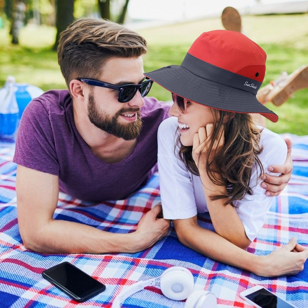 A couple enjoying a sunny picnic on a plaid blanket, wearing a stylish red and gray sun hat, with headphones and a smartphone nearby.