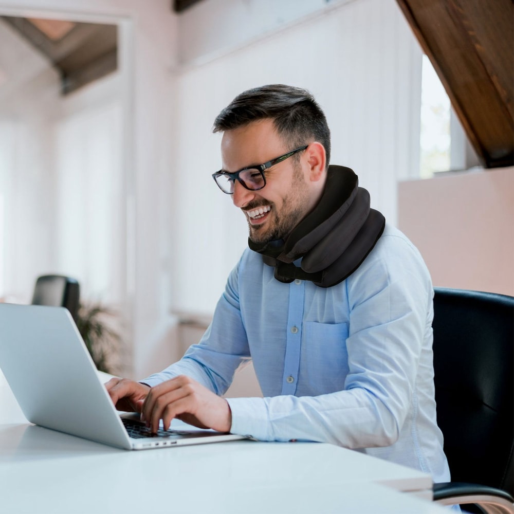 Person seated at a desk wearing a supportive neck pillow while working on a laptop in a modern office setting.