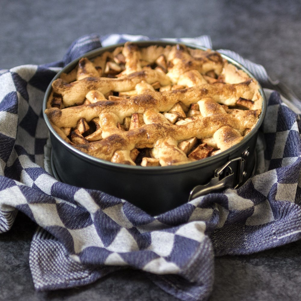 Freshly baked apple pie in a round springform pan, with a golden brown lattice crust, resting on a blue and white checkered kitchen towel.