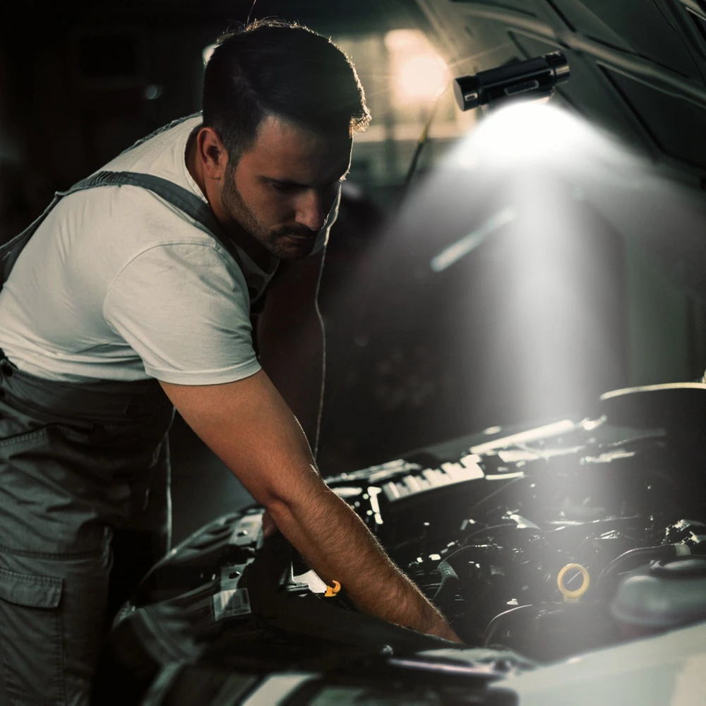 Mechanic working on a car engine with a focused light illuminating the area under the hood, emphasizing hands-on automotive repair expertise.