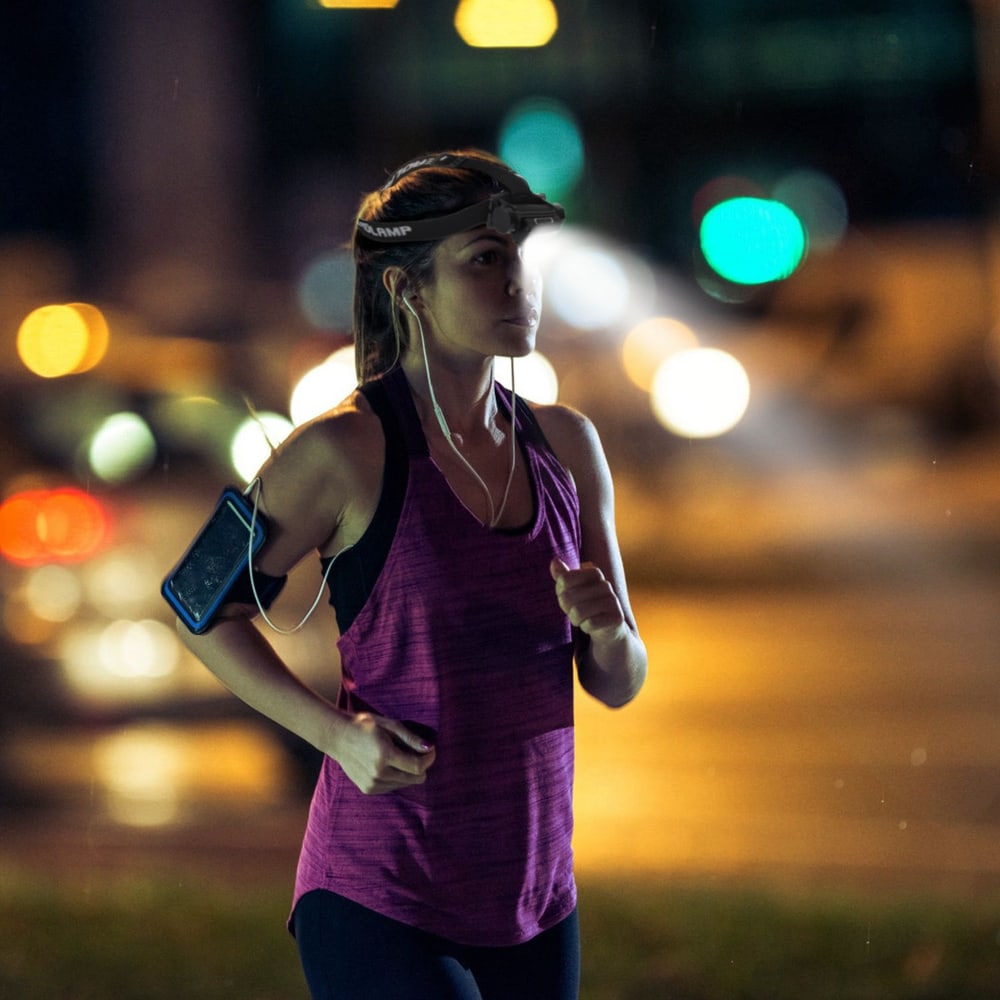 Woman jogging at night wearing a purple athletic tank top and headphones, with a smartphone armband, illuminated by city lights in the background.