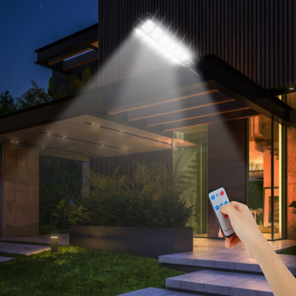 Remote-controlled outdoor LED floodlight illuminating a modern home entrance at night, with a hand holding the remote in the foreground.