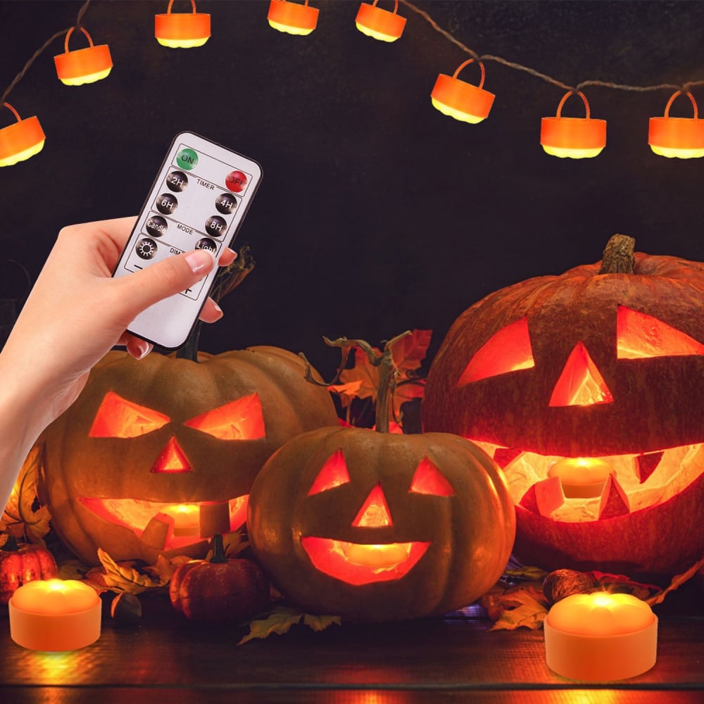 Remote-controlled LED Halloween pumpkin lights with a hand holding the remote, featuring carved pumpkins glowing warmly among autumn leaves and decorative candy-style lights in the background.