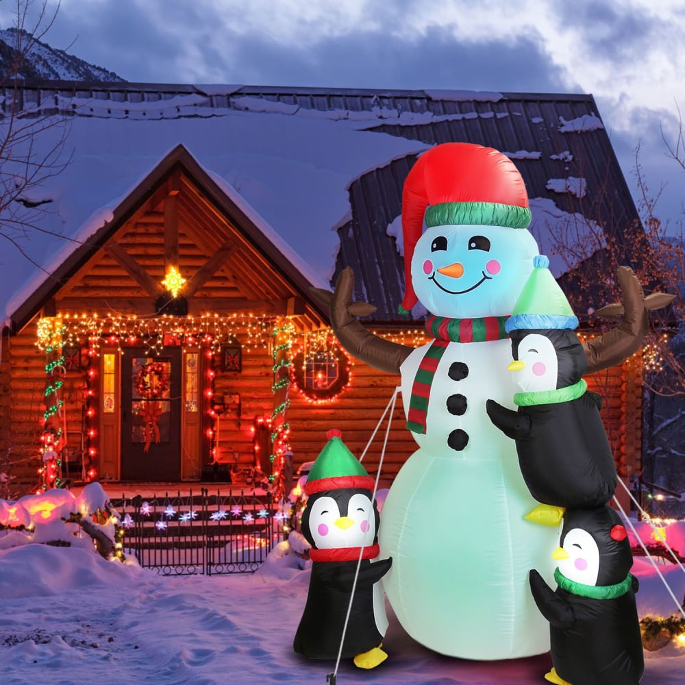Inflatable Christmas snowman with penguins in front of a cozy, decorated holiday cabin covered in snow.