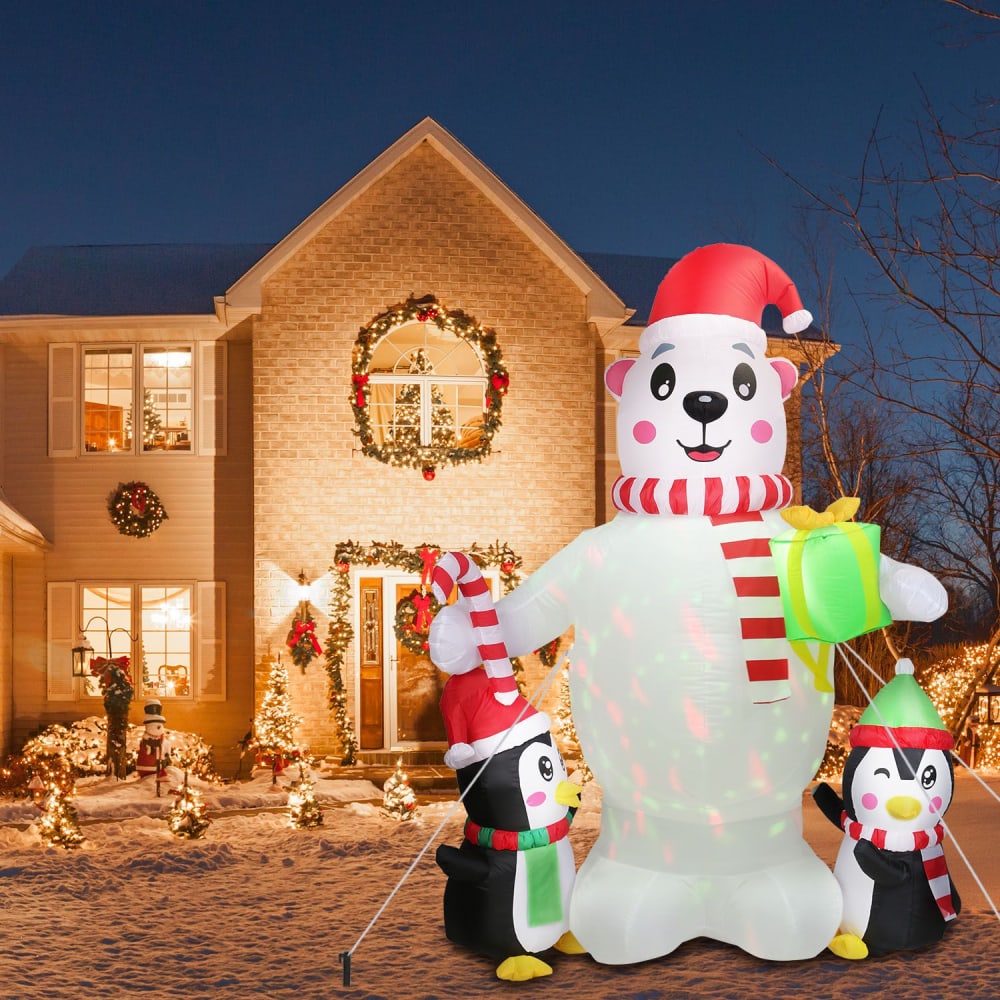 Inflatable Christmas decoration featuring a cheerful polar bear with a gift and two smiling penguins, set in front of a beautifully lit home adorned with holiday wreaths and twinkling lights.