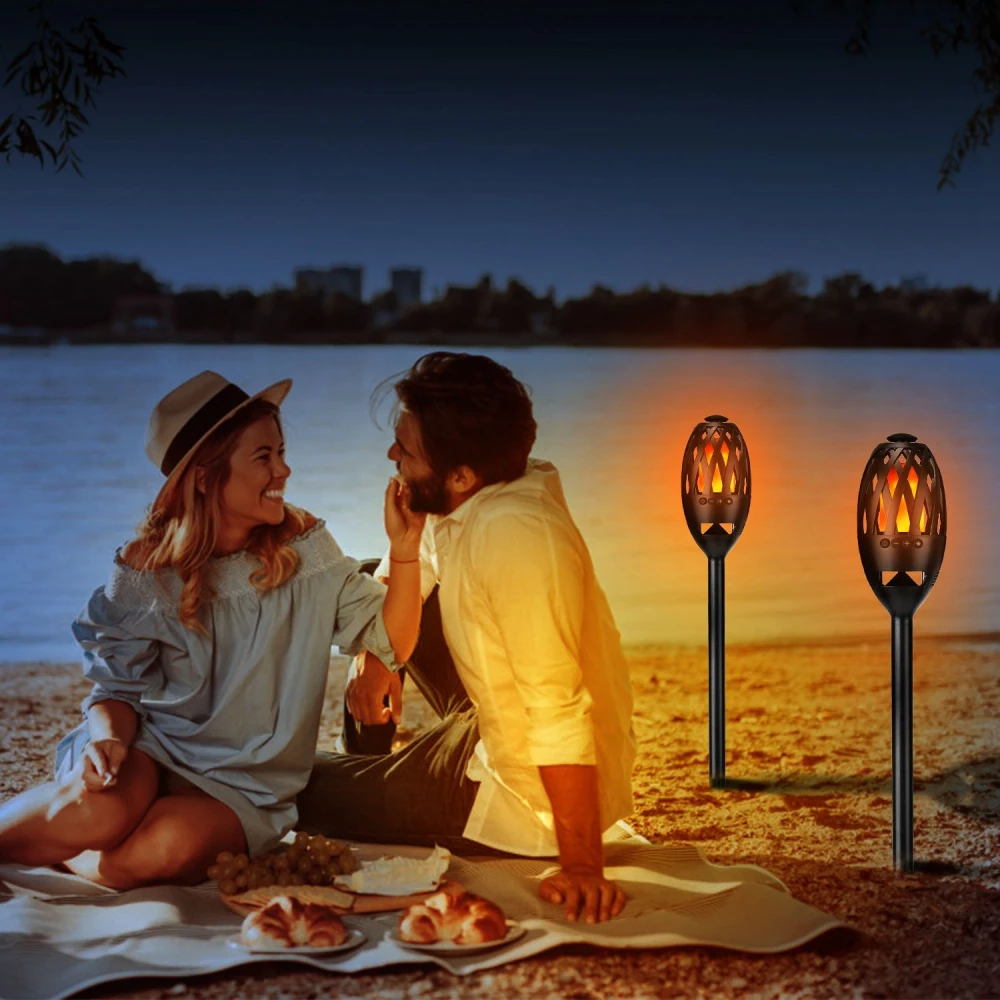 Couple enjoying a romantic picnic by the lake at sunset, featuring outdoor flame torches, soft lighting, and a spread of pastries and grapes on a blanket.