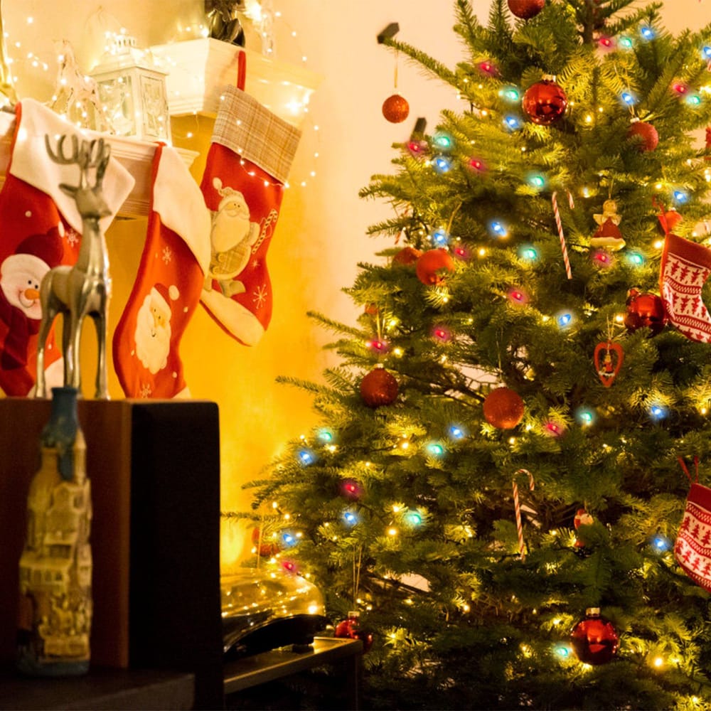 Festively decorated Christmas tree with colorful lights and ornaments, surrounded by red stockings and a silver reindeer statue, creating a warm holiday atmosphere.