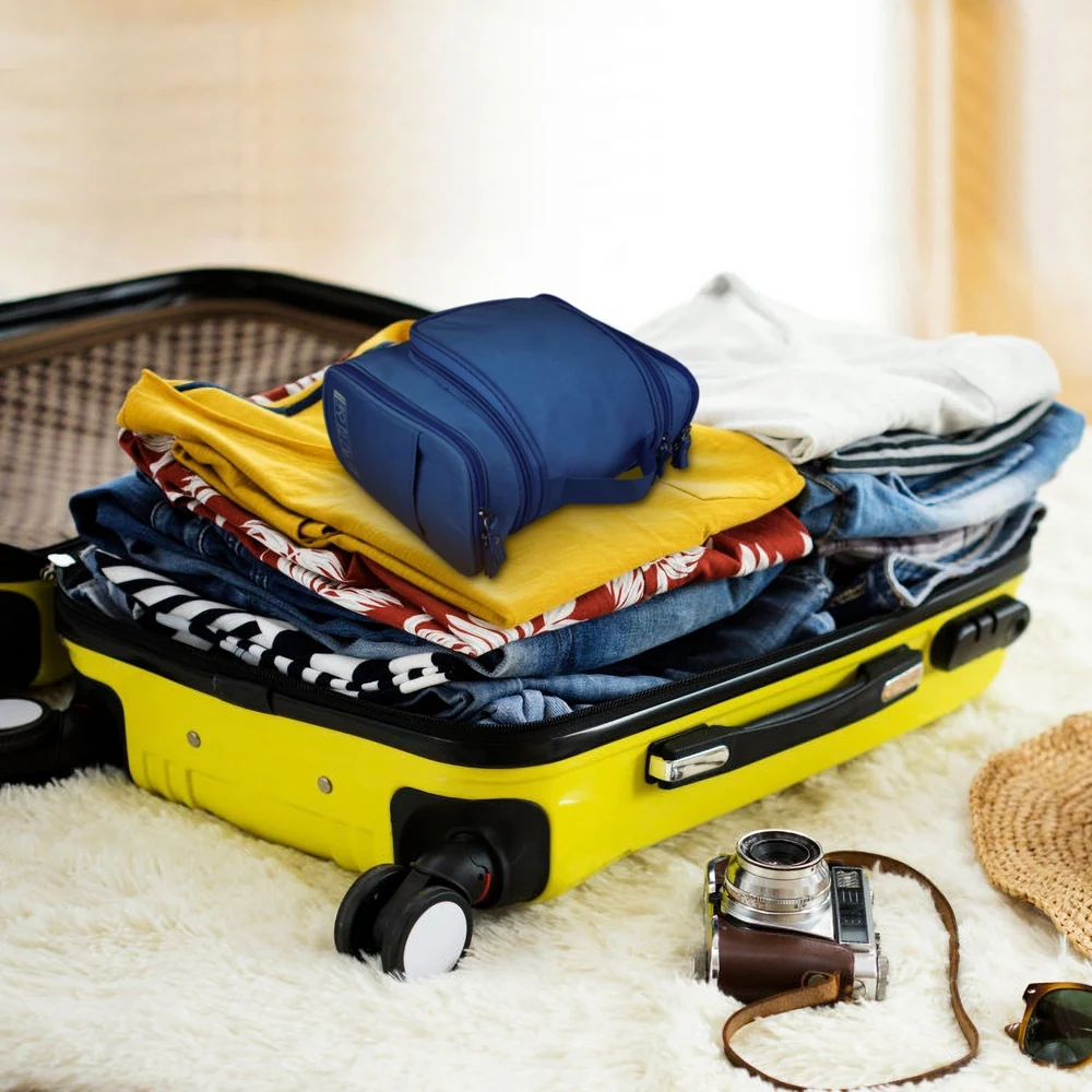 Compact navy travel organizer bag placed on neatly folded clothes inside a yellow suitcase, with a vintage camera and straw hat beside it.