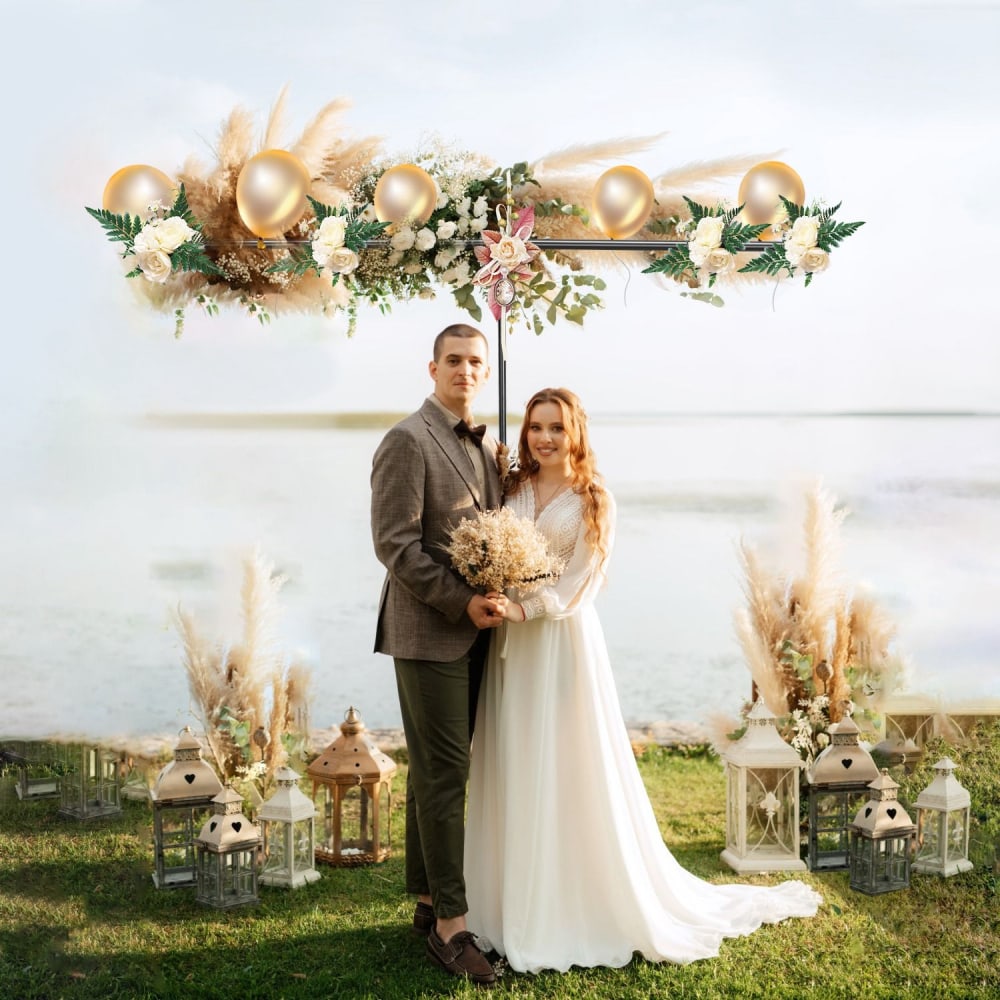 A bride and groom stand together in front of a beautifully decorated backdrop, featuring gold balloons, greenery, and floral arrangements by a serene body of water, surrounded by elegant lanterns and pampas grass.