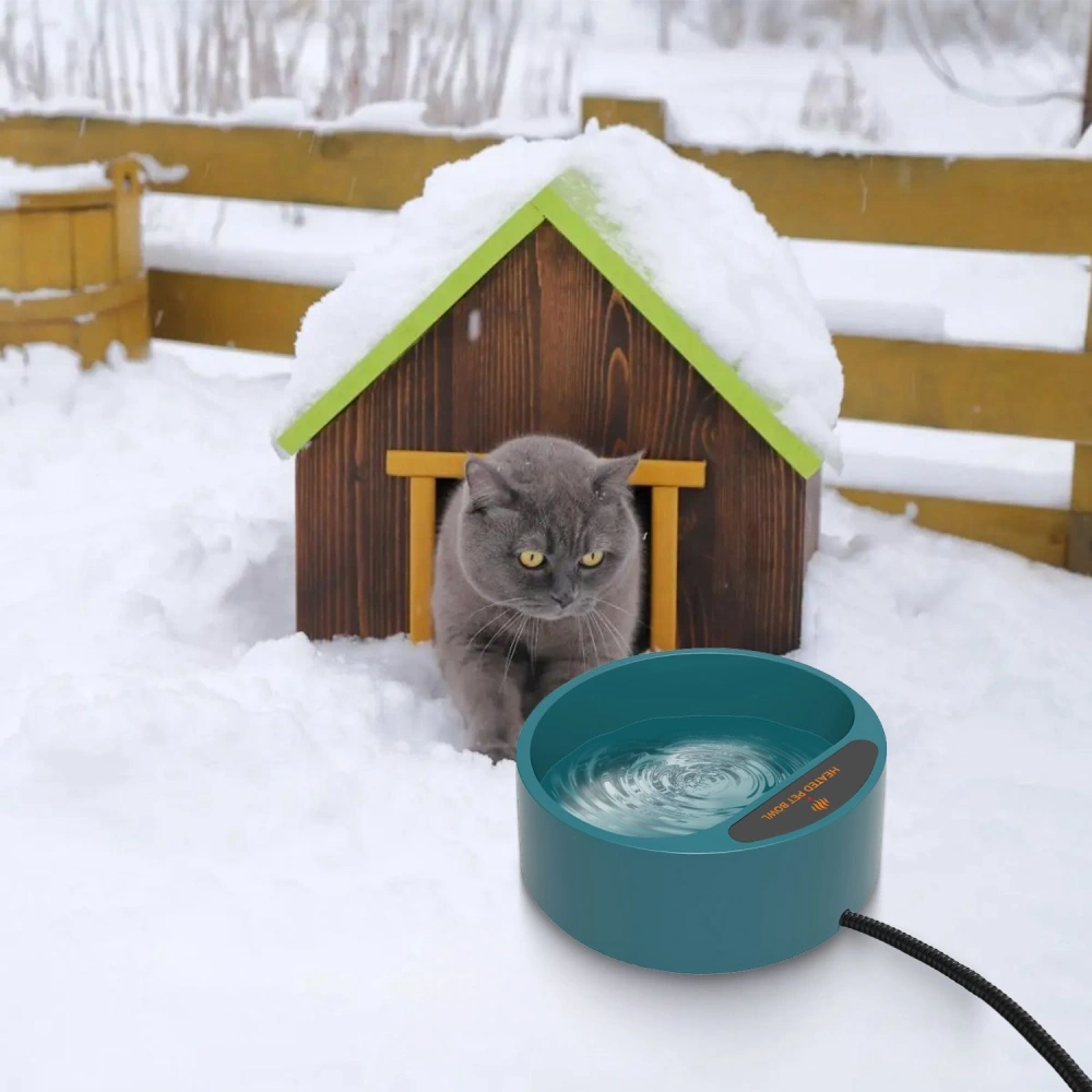 Gray cat approaching a heated water bowl in the snow, next to a cozy wooden cat house with a green roof.