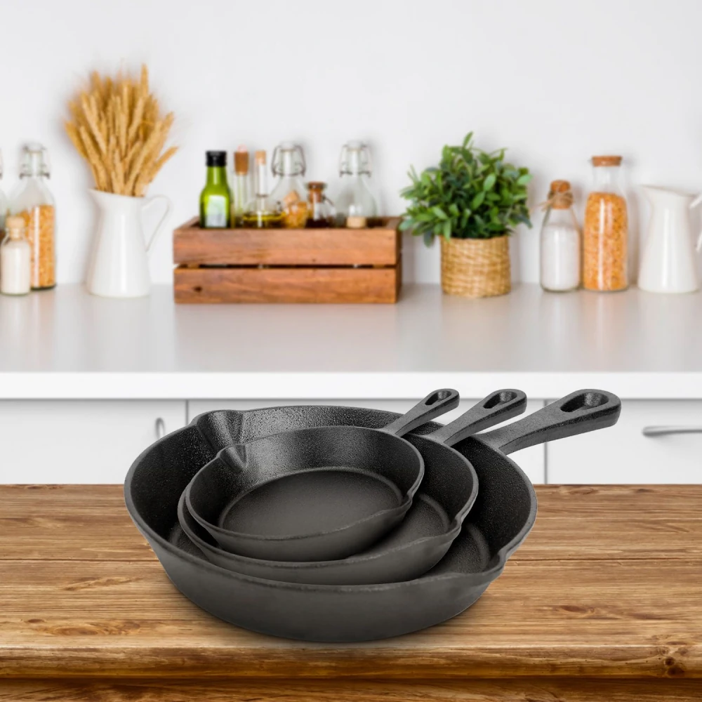 Three nested cast iron skillets displayed on a wooden countertop with a modern kitchen background, featuring a variety of glass bottles and a small potted plant.