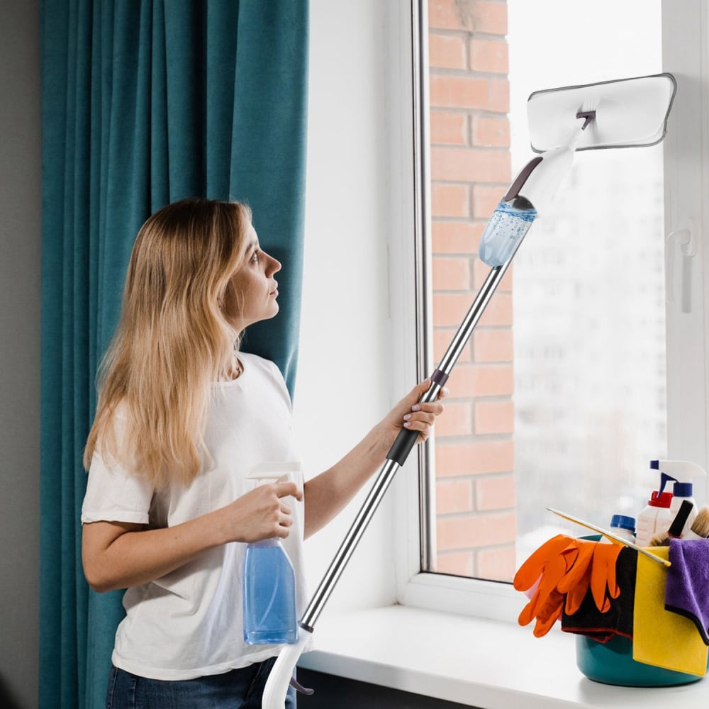 Woman cleaning a window with a spray bottle and a extendable window cleaner tool, surrounded by cleaning supplies including gloves and cloths, in a bright room with teal curtains.