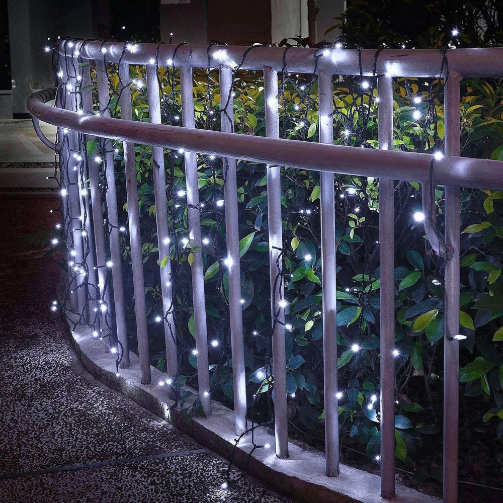 Outdoor string lights wrapped around a metal railing, illuminating nearby green foliage at night, creating a warm and festive ambiance.