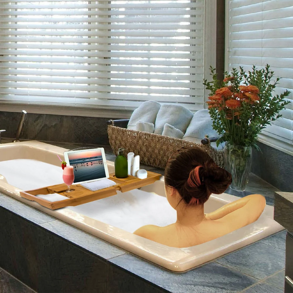 Woman relaxing in a luxurious bathtub with a wooden tray holding a tablet, a cocktail, and bath essentials, surrounded by soft towels and vibrant flowers, creating a serene spa-like atmosphere.