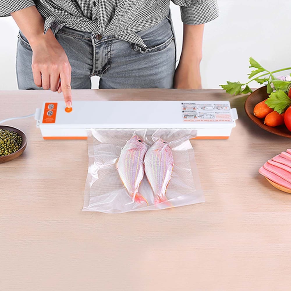 Person using a vacuum sealer to package two fish in a clear bag, with fresh vegetables and beans on a wooden countertop.